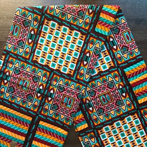 Op de afbeelding: Een kleurrijke stof met een geometrisch patroon in tinten rood, oranje, geel, blauw en wit. Het patroon bevat vierkanten, ruiten en andere vormen.