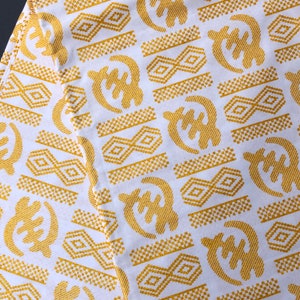 May include: White fabric with a repeating pattern of gold Adinkra symbols. The symbols include geometric shapes and stylized forms, creating a visually rich and textured design. The fabric is likely used for clothing or decorative purposes.