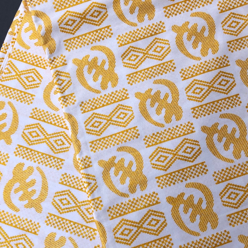 May include: White fabric with a repeating pattern of gold Adinkra symbols. The symbols include geometric shapes and stylized forms, creating a visually rich and textured design. The fabric is likely used for clothing or decorative purposes.
