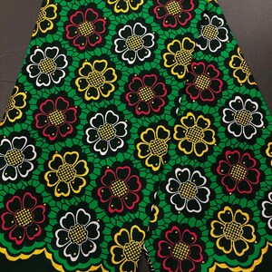 Puede incluir: Tela verde con un patrón repetido de flores negras, blancas, rojas y doradas. Las flores tienen un diseño geométrico y están delineadas en oro.