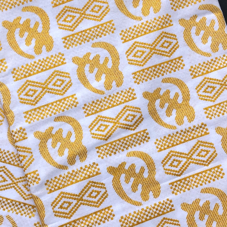 May include: White fabric with a repeating pattern of gold Adinkra symbols. The symbols include the Gye Nyame symbol, a symbol of the omnipotence of God, and other geometric shapes. The fabric is likely used for clothing or decorative purposes.