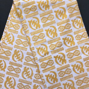 May include: White fabric with a repeating pattern of gold Adinkra symbols. The symbols include the Sankofa bird and other geometric shapes. The fabric is folded, showing the pattern's continuity. The fabric is likely used for clothing or decorative purposes.