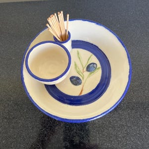 May include: Ceramic olive serving dish with a cream-colored base and a blue rim. The dish features a small bowl and a toothpick holder. The design includes a painted olive branch with blue olives.