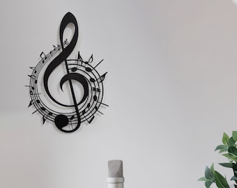 Op de afbeelding: Een zwarte metalen vioolsleutel wanddecoratie met muzieknoten eromheen. Een zilveren microfoon, koptelefoon, een laptop en een glas vloeistof staan op een houten tafel voor een witte muur.
