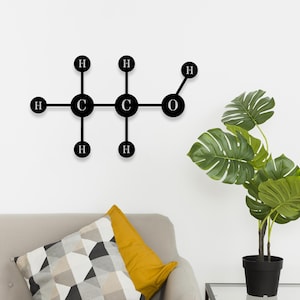 Op de afbeelding: Zwarte metalen wandkunst die een chemische formule met de letters H, C en O afbeeldt. De kunst is gemonteerd op een witte muur met een groene plant in een zwarte pot op de achtergrond.