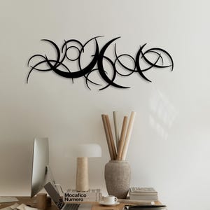 Könnte beinhalten: Schwarze Metall-Wandkunst mit einem Design aus miteinander verbundenen Halbmond- und Kreisformen. Das abstrakte Kunstwerk ist an einer weißen Wand über einem Schreibtisch mit Computer, Lampe und Dekorationsgegenständen angebracht. Thema: moderne Wohnkultur.