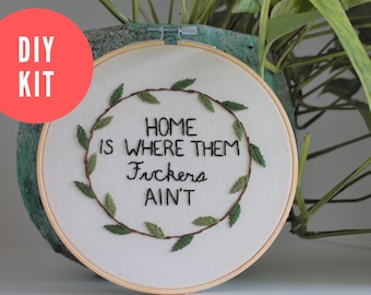 Puede incluir: Kit de bordado DIY con un aro blanco con una corona de hojas verdes y el texto "Home is where them F*ckers ain't".