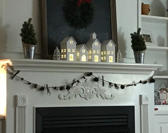 May include: A decorated fireplace mantel with a chalkboard, wreath, and illuminated miniature houses. Two small potted evergreen trees flank the chalkboard. A garland of pinecones and twigs adorns the mantel. The fireplace is black.