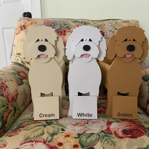May include: Three wooden dog-shaped planters in cream, white, and golden colors. Each has a black nose, pink tongue, and a small rectangular sign at the base with the color name. The planters are on a floral patterned chair.