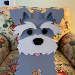 May include: A wooden Schnauzer dog decoration with a gray body, white beard and eyebrows, and black eyes. The dog has a pink tongue sticking out. The decoration is placed in front of a floral patterned chair.