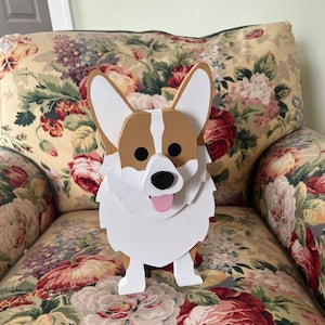 May include: A wooden corgi dog figurine with a white, brown, and black color scheme. The dog has large ears, a black nose, and a pink tongue sticking out. The figurine is sitting on a floral patterned chair.