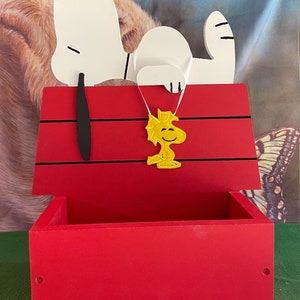 May include: A red wooden box shaped like Snoopy's doghouse, with Snoopy lying on top. A yellow Woodstock figure hangs from the doghouse. The box has a rectangular opening and is painted red with black accents.