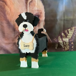 May include: A wooden dog-shaped planter painted in black, white, and tan. The dog has a bone-shaped tag that says "Lucy". The planter is designed to hold small plants or flowers, adding a touch of whimsy to any garden or indoor space.