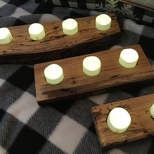 May include: Three rustic wooden candle holders, each with three glowing white LED candles. The candle holders are made from rough-hewn wood, showcasing the natural grain and texture. The arrangement is on a black and white checkered blanket.