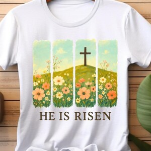 Puede incluir: Camiseta blanca con un gráfico de una cruz en una colina con flores. El texto "He is Risen" está impreso debajo del gráfico.