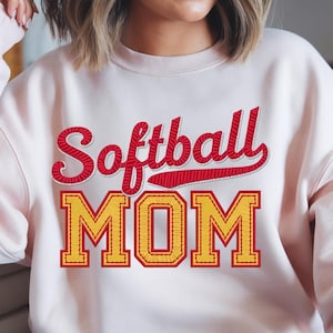 Könnte beinhalten: Ein rosa Sweatshirt mit dem Text "Softball MOM" in roten und gelben Stickbuchstaben.