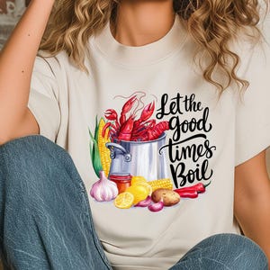 May include: A white t-shirt with a graphic design of a pot of crawfish, corn on the cob, lemons, garlic, and red peppers. The text "Let the good times Boil" is written in black cursive lettering.