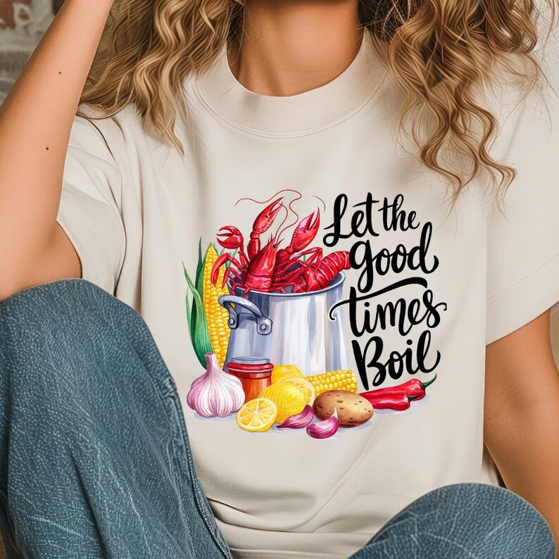 May include: A white t-shirt with a graphic design of a pot of crawfish, corn on the cob, lemons, garlic, and red peppers. The text "Let the good times Boil" is written in black cursive lettering.