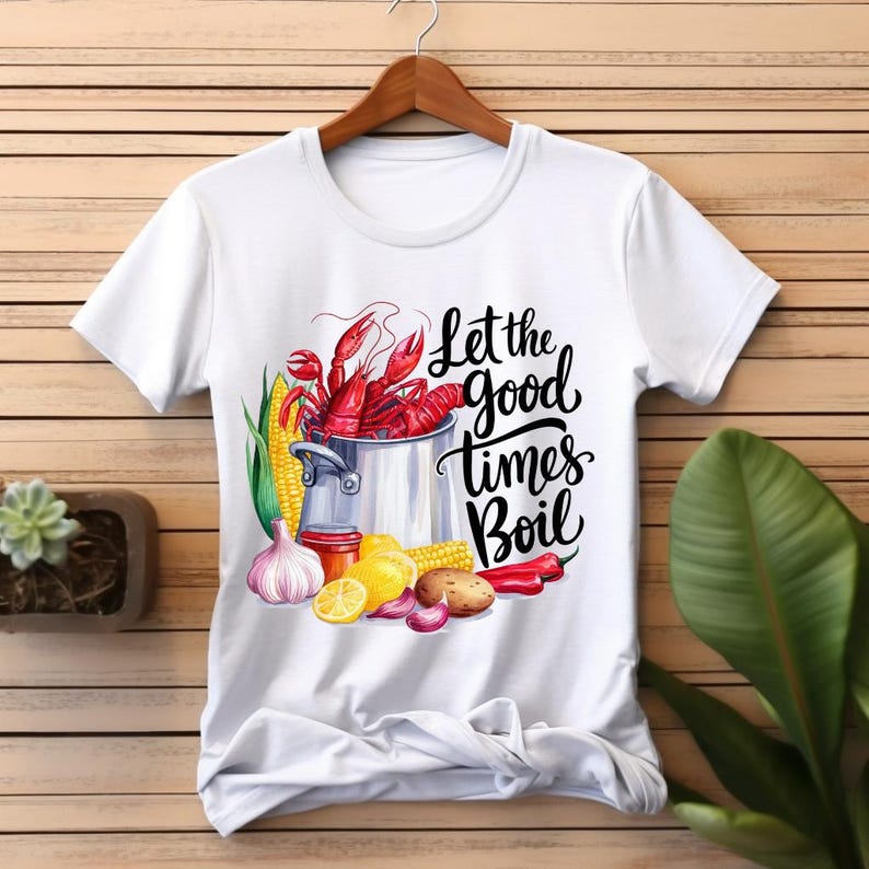 May include: White t-shirt with a graphic of a pot of crawfish, corn, garlic, lemons, potatoes, and red peppers. The text "Let the good times boil" is printed on the shirt.