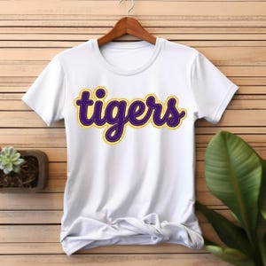 May include: A white short-sleeved t-shirt featuring the word "tigers" printed in a cursive font across the chest. The text is dark purple with a thick yellow outline and a stitched effect, displayed on a plain white fabric background.