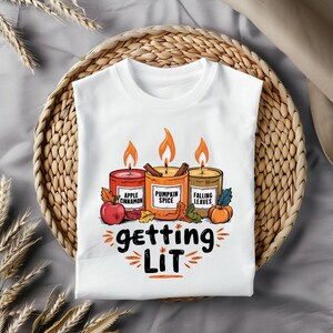 Puede incluir: Camiseta blanca con un gráfico de tres velas encendidas con los aromas Manzana Canela, Especias de Calabaza y Hojas Caídas. La frase "getting LIT" está escrita debajo. La camiseta está sobre una bandeja tejida.