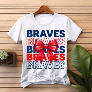 Op de afbeelding: Een wit t-shirt met korte mouwen waarop het woord BRAVES vijf keer is gedrukt in afwisselend effen marineblauw, marineblauwe omlijning en effen rood, gecentreerd met een grote, glanzende rode strik over de middelste tekst heen.