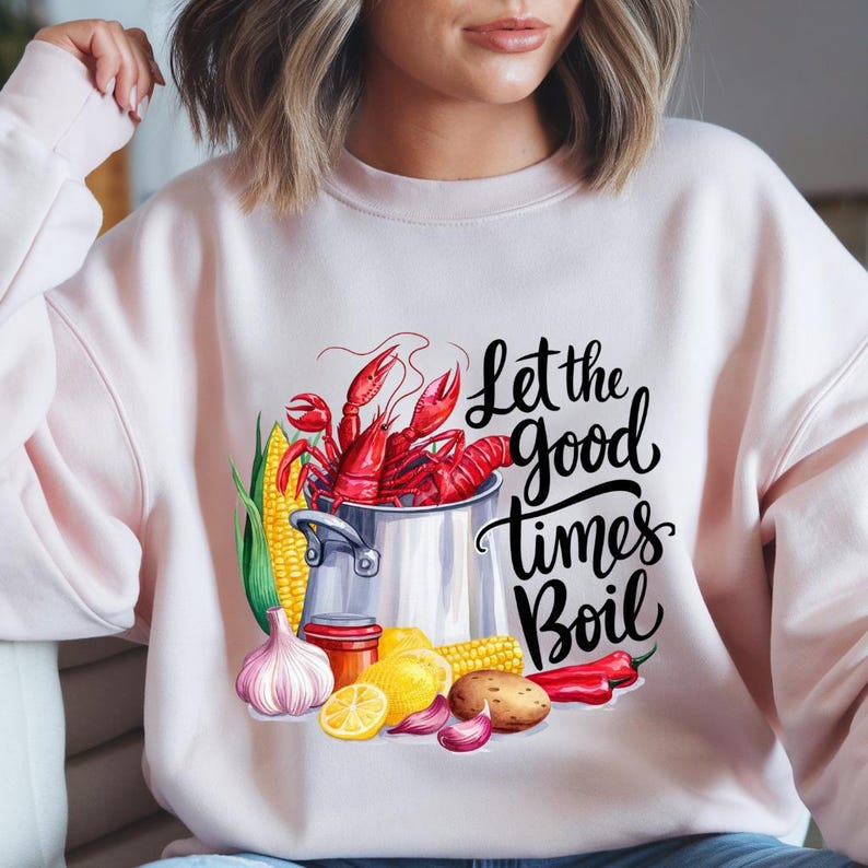 May include: A pink sweatshirt with a graphic design of a pot of crawfish, corn, lemons, garlic, and red peppers. The text "Let the good times boil" is written in black cursive lettering.