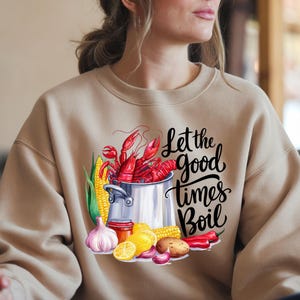 May include: A tan sweatshirt with a graphic of a pot of crawfish, corn on the cob, garlic, lemons, and red peppers. The text "Let the good times boil" is printed in black on the sweatshirt.
