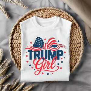 Puede incluir: Camiseta blanca con un lazo rojo, blanco y azul con estrellas y el texto "TRUMP Girl" en rojo y azul.