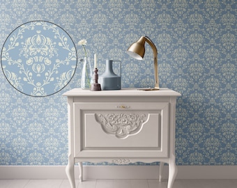May include: A white nightstand with ornate carvings and curved legs stands against a blue wall with a repeating floral pattern. A gold desk lamp, a vase, and a candle holder are on the nightstand. A close-up of the wallpaper pattern is in a circle.