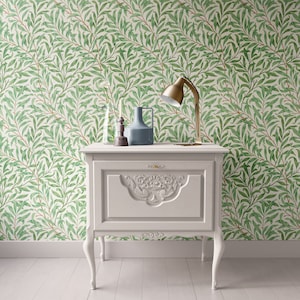 May include: A white bedside table with a carved design on the front drawer. The bedside table has three legs and is positioned in front of a green and white wallpaper with a floral pattern. A gold lamp, a clear glass vase with dried flowers, a black candle holder, and a blue ceramic vase are on top of the bedside table.