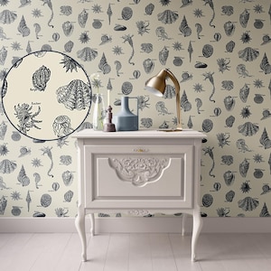 Puede incluir: Una mesita de noche blanca con un diseño tallado en la parte delantera. La mesita de noche tiene una lámpara con una base dorada y una pantalla blanca, un jarrón azul y una vela en la parte superior. La pared detrás de la mesita de noche está cubierta con un papel tapiz con un patrón en blanco y negro de conchas marinas y otras formas de vida marina.