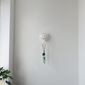 May include: A white, wall-mounted decorative hook in the shape of a stylized owl's head. A black lanyard with a collection of keys and decorative charms hangs from the hook. The background is a plain white wall.