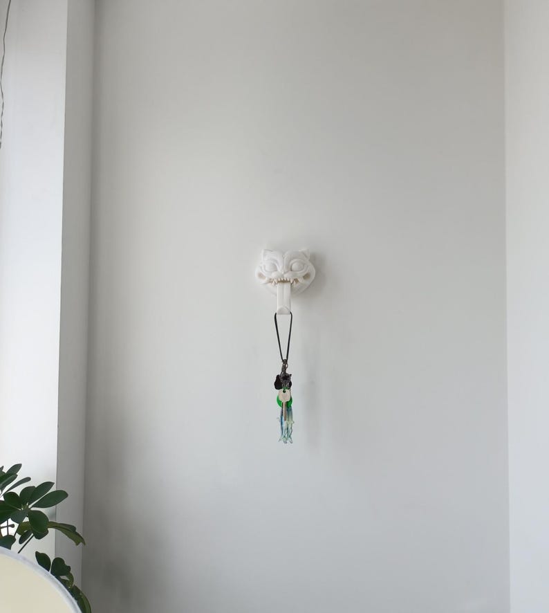 May include: A white, wall-mounted decorative hook in the shape of a stylized owl's head. A black lanyard with a collection of keys and decorative charms hangs from the hook. The background is a plain white wall.