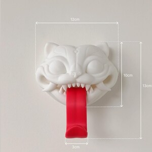 May include: A white cat-shaped wall hook with a red tongue, measuring 12cm wide, 10cm high, and 3cm deep. The hook is 5 inches tall. The cat has sharp teeth and is mounted on a white wall. The overall height is 13cm.