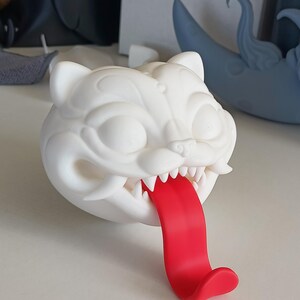 May include: A white, stylized cat head sculpture with a red tongue extending out. The sculpture has sharp teeth and detailed facial features. The cat head is sitting on a white surface.