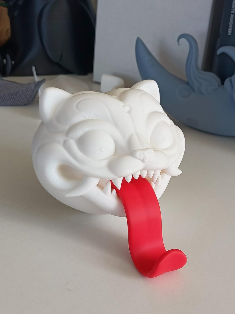 May include: A white, stylized cat head sculpture with a red tongue extending out. The sculpture has sharp teeth and detailed facial features. The cat head is sitting on a white surface.
