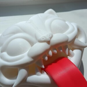 May include: A white, stylized cat head sculpture with sharp teeth and a protruding red tongue. The sculpture has large, expressive eyes and is likely made of a 3D-printed material.