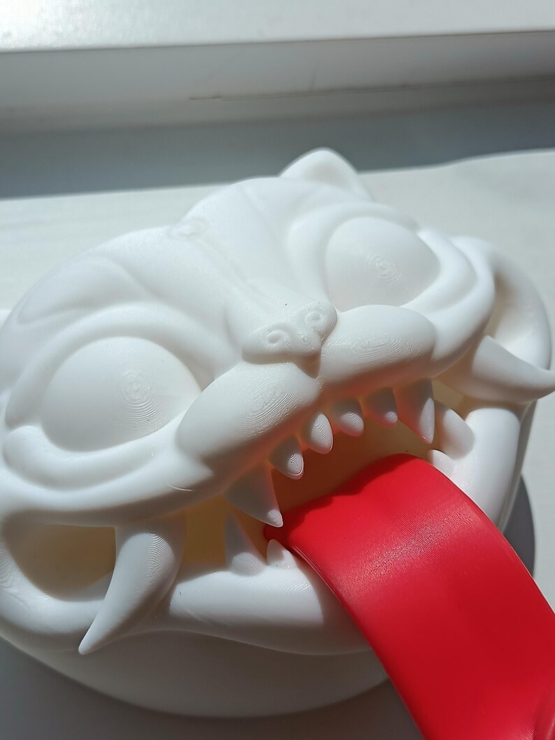 May include: A white, stylized cat head sculpture with sharp teeth and a protruding red tongue. The sculpture has large, expressive eyes and is likely made of a 3D-printed material.