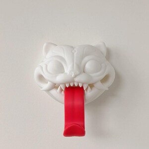 May include: A white cat-shaped wall hook with a red tongue. The cat has sharp teeth and is mounted on a white wall. The hook is designed to hold items.