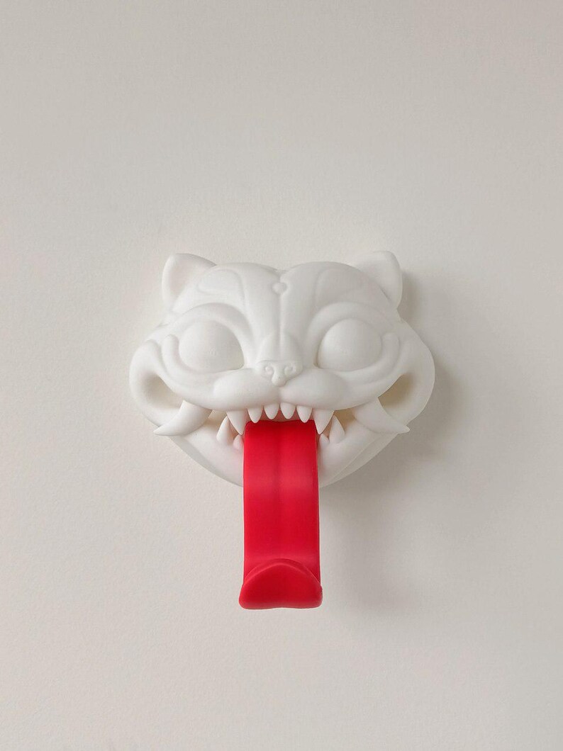 May include: A white cat-shaped wall hook with a red tongue. The cat has sharp teeth and is mounted on a white wall. The hook is designed to hold items.
