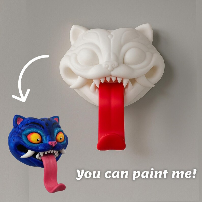 May include: A white cat-shaped wall hook with a red tongue, and a blue cat head with a pink tongue. The text "You can paint me!" is at the bottom. The hook is designed to hold items.