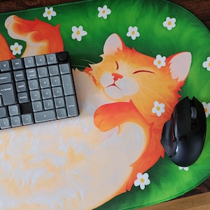 May include: A large oval desk mat featuring a vibrant green background with white flowers and a large orange cat sleeping on its back. A grey mechanical number pad and a black computer mouse are placed on top of the mat surface.