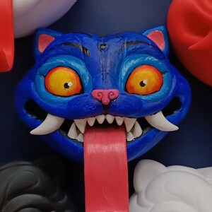 May include: A blue cat-faced sculpture with a long red tongue, sharp white teeth, and yellow eyes. The cat has pink accents on its ears and nose. Other cat faces in white, black, and red are visible in the background.