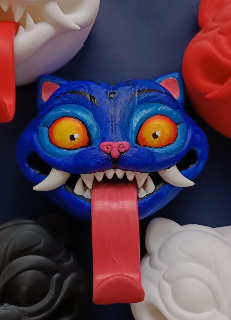 May include: A blue cat-faced sculpture with a long red tongue, sharp white teeth, and yellow eyes. The cat has pink accents on its ears and nose. Other cat faces in white, black, and red are visible in the background.