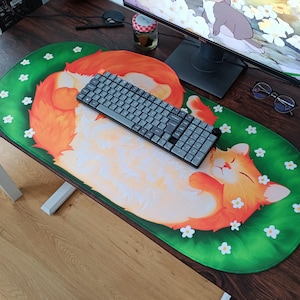 May include: An oval desk mat featuring a large, sleeping orange cat with white fur on its chest and belly, resting on a vibrant green background decorated with small white flowers. A dark grey mechanical keyboard sits on top of the mat.