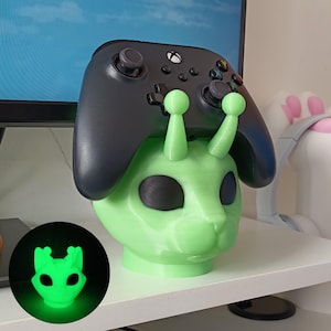 Può includere: Un supporto per controller a forma di gatto alieno verde con un controller Xbox nero sopra. Il supporto ha grandi occhi neri e due sporgenze simili ad antenne. Una versione più piccola e luminosa del supporto è nell'angolo in basso a sinistra.