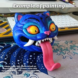May include: A blue, stylized tiger head sculpture with a long pink tongue, white fangs, and large yellow eyes. The text "Example of painting" is at the top. The text at the bottom says the original color is white.