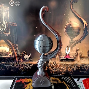 May include: A silver trophy with a spherical top and curved design, mounted on a dark wooden base. The trophy is positioned in front of a computer screen displaying a video game scene with a character in a red cloak.
