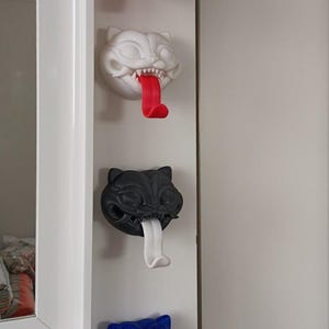 May include: Four cat-shaped wall hooks in red, white, black, and blue. Each hook has a protruding tongue that serves as the hook. The cat faces have sharp teeth and are mounted on a white wall.